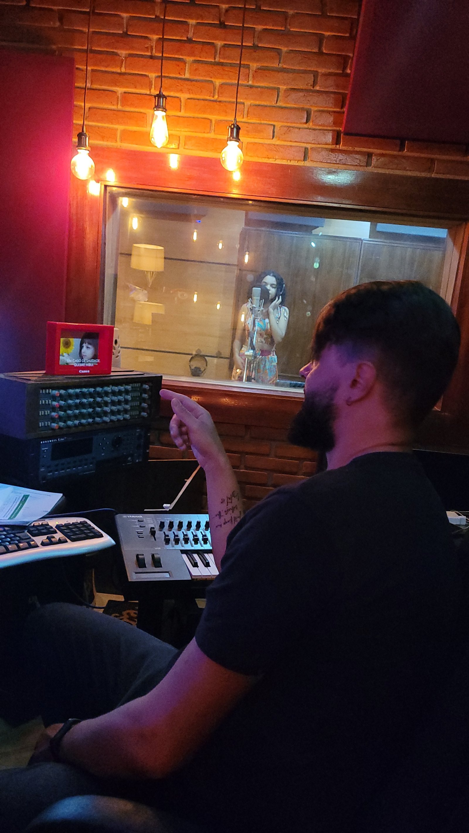 Thiago Melo dirigindo uma gravação no estúdio, ao teclado, com a cantora na cabine